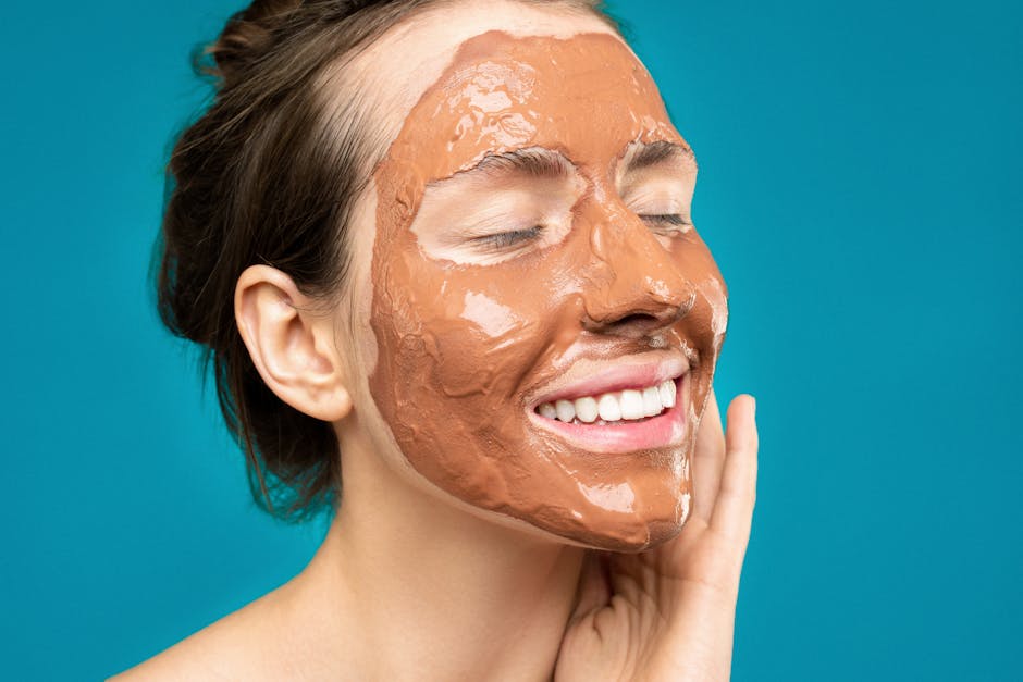 Close-up of a woman smiling with a clay mud mask on her face on a turquoise background.